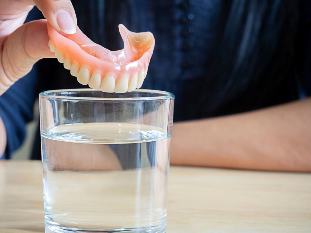 Fingers placing upper arch of dentures into glass of clear fluid